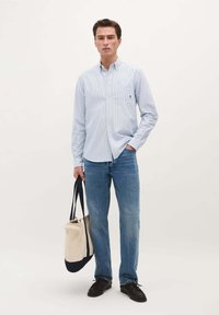 Lichtblauw gestreept overhemd met knopen en een zak, gecombineerd met een blauwe spijkerbroek en zwarte schoenen. Houdt een beige en marineblauwe boodschappentas vast.