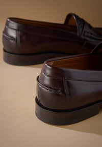 Dunkelbraune Lederloafers mit einer glatten Oberfläche, runder Zehenform und niedrigem Gummiabsatz. Mit Steppnähten und einem eleganten Design.