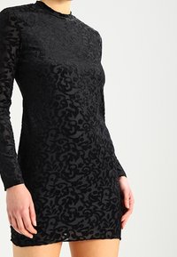 Abito nero a maniche lunghe realizzato in tessuto testurizzato con un motivo floreale. Collo alto e silhouette aderente, che termina sopra il ginocchio.