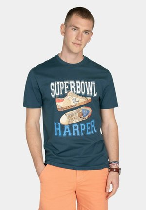 Młody mężczyzna w ciemnoturkusowej koszulce "Superbowl Harper" z grafiką sneakersów i pomarańczowych szortach, stojący z jedną ręką w kieszeni.