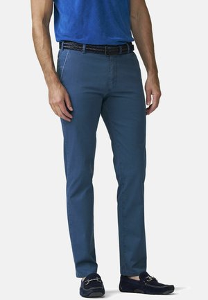 Man draagt blauwe slim-fit broek, zwarte gevlochten riem, marineblauwe loafers en een blauw t-shirt in de broek gestopt, staand tegen een neutrale achtergrond.