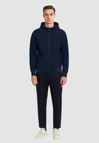Felpa con zip navy dal tessuto strutturato, con tasca frontale e cappuccio regolabile, abbinata a pantaloni neri e sneakers bianche.