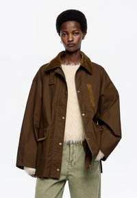 Hnedá, prehnaná bunda s kordovým golierom, cvokovými gombíkmi a bočnými vreckami. Nosená cez huňatý biely sveter a svetlozelené nohavice.