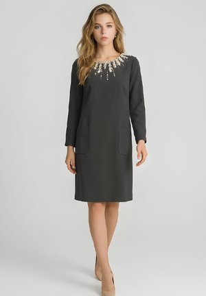 Joven mujer con un vestido negro hasta la rodilla, de manga larga y adornos de perlas alrededor del escote, lleva tacones de color nude.
