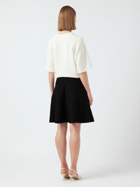 Femme aux cheveux bruns courts portant un pull blanc à manches courtes, une jupe noire jusqu'aux genoux et des escarpins à bride arrière blancs, se tenant de dos.