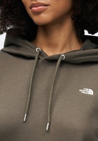 Φούτερ σε ελιά πράσινο με κουκούλα, με ασημένιες οπές και κορδόνια. Στο μπροστινό μέρος εμφανίζεται λευκό λογότυπο στην αριστερή πλευρά. Ομαλή υφή.