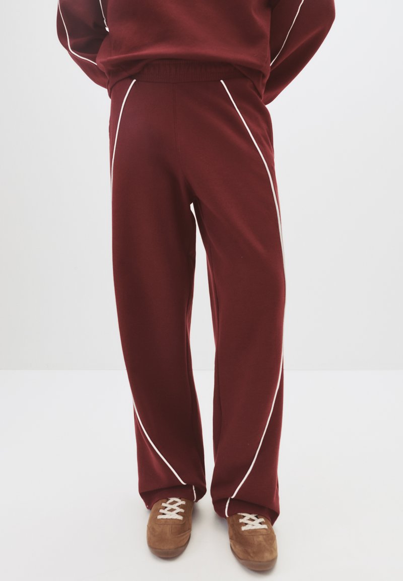 Person in burgunderfarbenen Jogginghosen mit weißen Paspeln und braunen Sneakers, die vor einem schlichten weißen Hintergrund stehen.