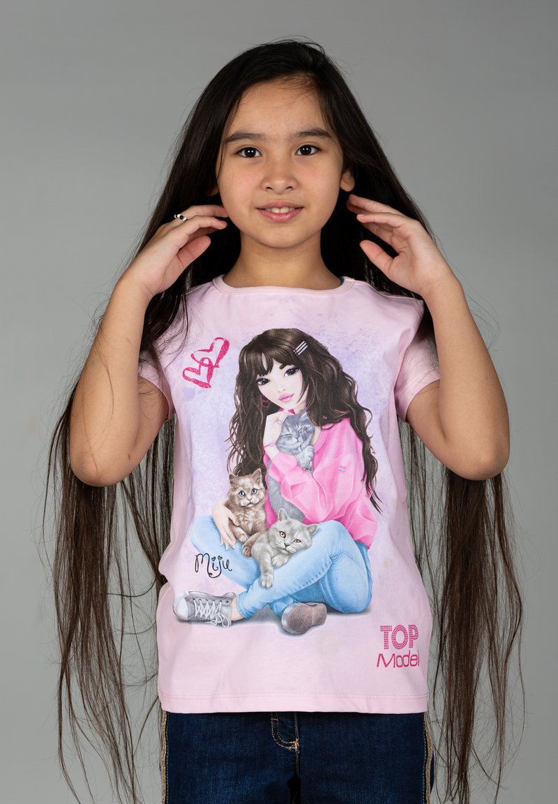 T-shirt a maniche corte di colore rosa chiaro con una grafica che raffigura una ragazza con lunghi capelli scuri che tiene in braccio tre gattini, abbinato a jeans blu e sneakers.