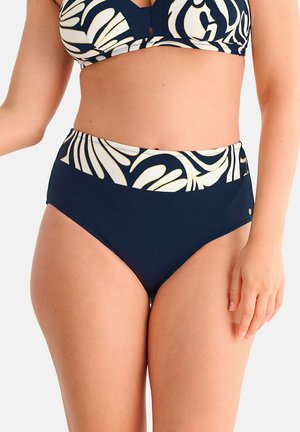 Femme portant un bas de bikini bleu marine à taille haute avec un motif de feuilles tropicales beige sur la ceinture.