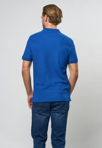 Muž stojící zády, oblečený v jasně modré polokošili s krátkými rukávy a tmavě modrých džínách na jednobarevném světlém pozadí.