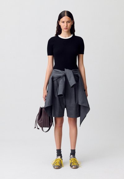 Mujer vestida con una camiseta negra ajustada de mangas cortas, pantalones cortos grises con una chaqueta a juego atada a la cintura, calcetines grises, zapatillas metálicas con cordones amarillos, sosteniendo un bolso marrón.