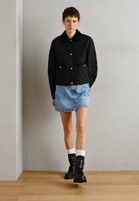 Chaqueta negra acolchada con botones de presión, falda de mezclilla azul claro y botas negras con calcetines blancos, de pie sobre un suelo de madera contra una pared gris.