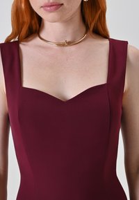 Bordeaux gekleed jurk met een sweetheart halslijn en brede bandjes. Glad stofoppervlak met minimale naden en strakke lijnen. Gouden chokeraccessoire.