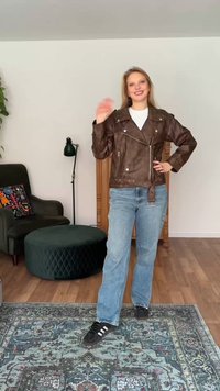 Tânără purtând o jachetă de piele maro, blugi albaștri şi adidaşi negri, zâmbitoare şi făcând cu mâna într-un living cu fotoliu și otomană.