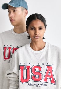 Felpa grigia con logo rosso "USA" e testo "NEW YORK", dotata di collo a giro e polsini a costine. Un cappellino in denim indossato da una figura sullo sfondo.
