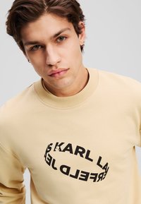 Sweatshirt beige clair avec un design de texte logo noir rond. Possède un col ras du cou et des poignets côtelés. Tissu doux avec une texture lisse.
