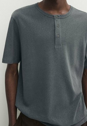 Homme portant un t-shirt henley à manches courtes gris foncé avec trois boutons sur le plastron avant.