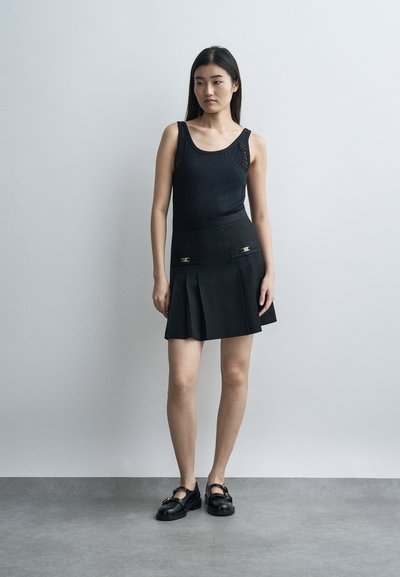 Γυναίκα που στέκεται φορώντας μαύρο αμάνικο τοπ, μαύρη πλισέ φούστα με διακοσμητικά κουμπιά και μαύρα μοκασίνια, μπροστά σε απλό γκρι φόντο.