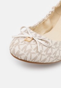 Ballerine beige à motif avec bordure froncée, un nœud blanc et un petit charme carré doré sur l\'orteil.