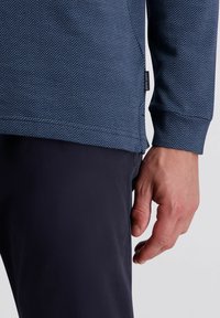Langärmliges Oberteil in Marineblau mit Fischgrätenmuster, gerade geschnittenem Saum und gerippten Bündchen. Getragen mit dunklen Hosen, wobei ein Teil einer Hand zu sehen ist.