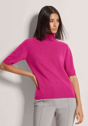 Femme aux cheveux bruns lisses portant un pull à col roulé rose vif à manches courtes et un pantalon gris clair, debout devant un fond clair.