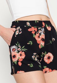 Femme portant un short noir à taille élastique avec un imprimé floral orange et blanc, main dans la poche, assorti à un top blanc court.