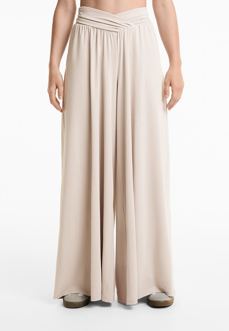 Beige weit geschnittene Hosen mit einem gerafften Taillenbund, aus einem weichen, dehnbaren Stoff, mit einem fließenden Design und einer glatten Textur.