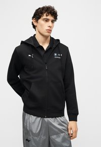 Νεαρός άνδρας που φοράει μαύρο φούτερ με φερμουάρ BMW Motorsport και γυαλιστ ερό γκρι αθλητικό παντελόνι, στέκεται μπροστά σε απλό φωτεινό φόντο.