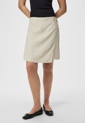 Γυναίκα που φοράει μπεζ κρεπ φούστα μέχρι το γόνατο, μαύρο αμάνικο τοπ και μαύρες μπαλαρίνες, στέκεται μπροστά σε απλό λευκό φόντο.