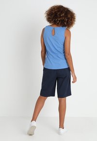 Blaues ärmelloses Top mit einem Schlüssellochdetail am Rücken, kombiniert mit marineblauen Shorts. Das Model trägt weiße Sneaker.