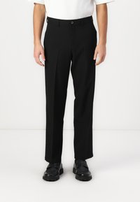 Μαύρο παντελόνι με κοπή ίσια, διαθέτει μια μπροστινή πτυχία, κούμπωμα με κουμπί και απαλό ύφασμα, συνδυασμένο με μαύρα παπούτσια.