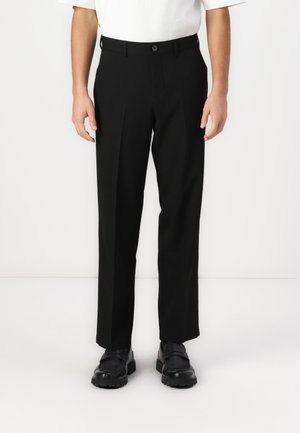 Schwarze Hose, taillierte Passform mit vorderen Falten, Knopf am Bund und geradem Beinansatz, kombiniert mit schwarzen Schuhen.