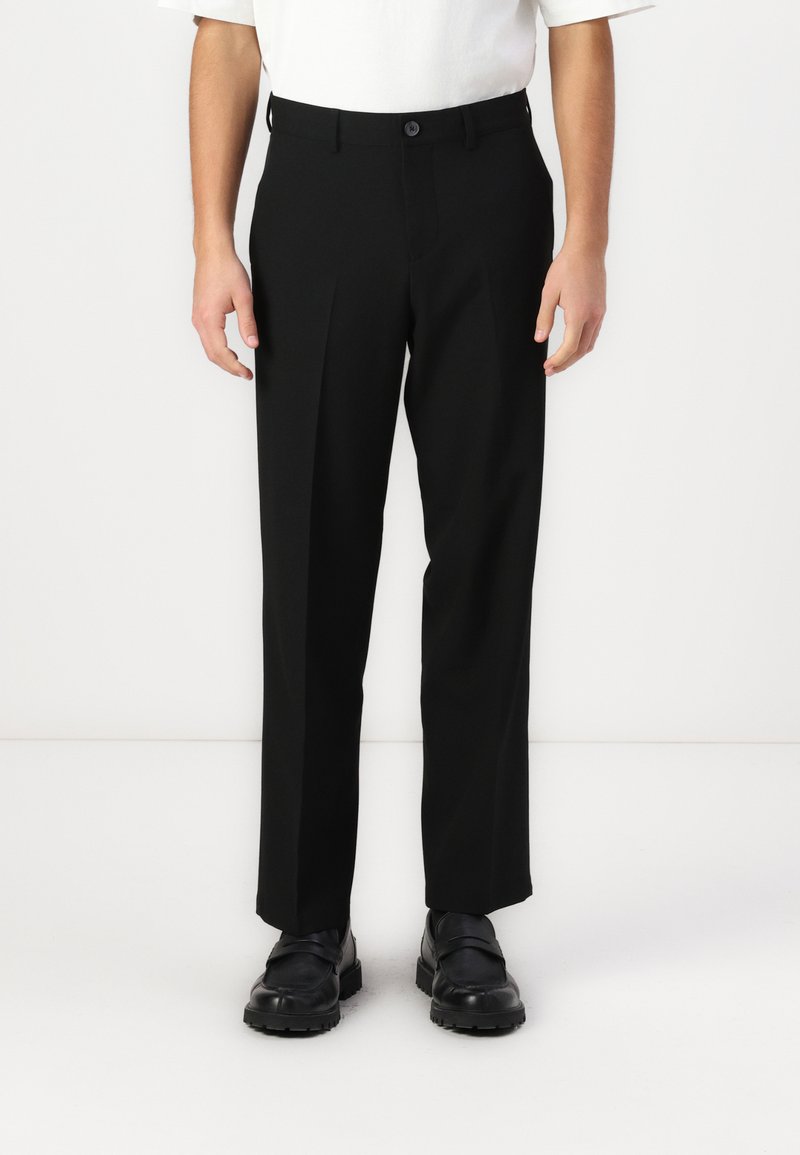 Μαύρο παντελόνι με κοπή ίσια, διαθέτει μια μπροστινή πτυχία, κούμπωμα με κουμπί και απαλό ύφασμα, συνδυασμένο με μαύρα παπούτσια.
