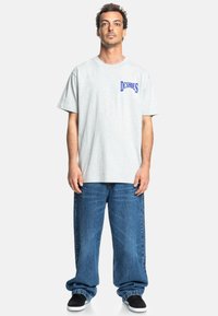 Gray cotton t-shirt with short sleeves and a blue "DC Shoes" logo on the left chest, paired with loose-fitting blue denim jeans and black sneakers.