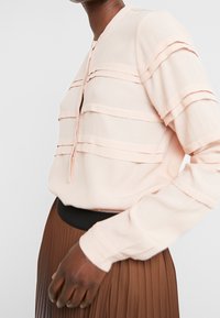 Persona che indossa una blusa rosa chiaro con pieghe orizzontali e una gonna plissettata marrone con una vita nera, su uno sfondo semplice.
