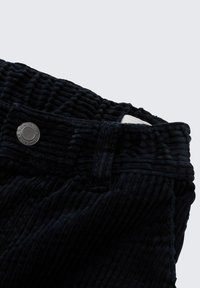 Pantalon en velours côtelé noir avec une surface texturée à côtes, doté d'une fermeture à bouton métallique et d'une étiquette en tissu apparaissant sur la ceinture.