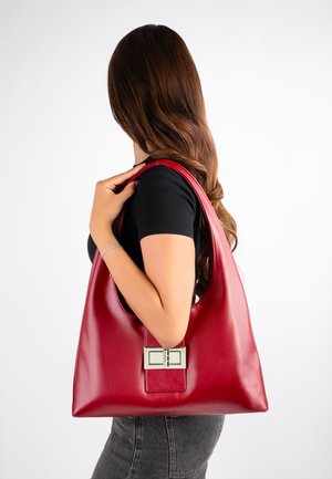 Femme aux longs cheveux bruns portant un haut noir et un jean gris, portant un grand sac à main en cuir rouge avec une boucle argentée sur son épaule.