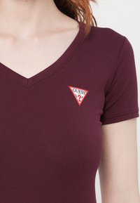 Inquadratura ravvicinata di un torso che indossa una maglietta V-neck bordeaux con un piccolo logo triangolare rosso e bianco di Guess sul lato sinistro del petto.
