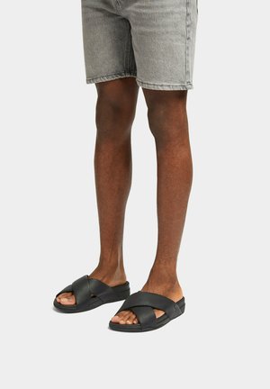 Man die lichtgrijze denim shorts en zwarte slides met kruisbandjes draagt, staande op een effen lichtgrijze achtergrond.