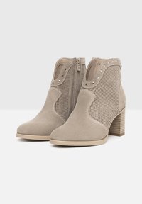 Bottine en suède beige avec un panneau en maille texturé, fermeture éclair sur le côté et clous argentés autour de l'encolure. Talon bloc avec une finition claire ressemblant à du bois.