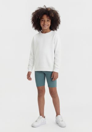 Kind mit lockigem Haar, das einen weißen langärmligen Pullover, türkisfarbene Radlerhosen und weiße Turnschuhe trägt, steht lächelnd vor einem weißen Hintergrund.