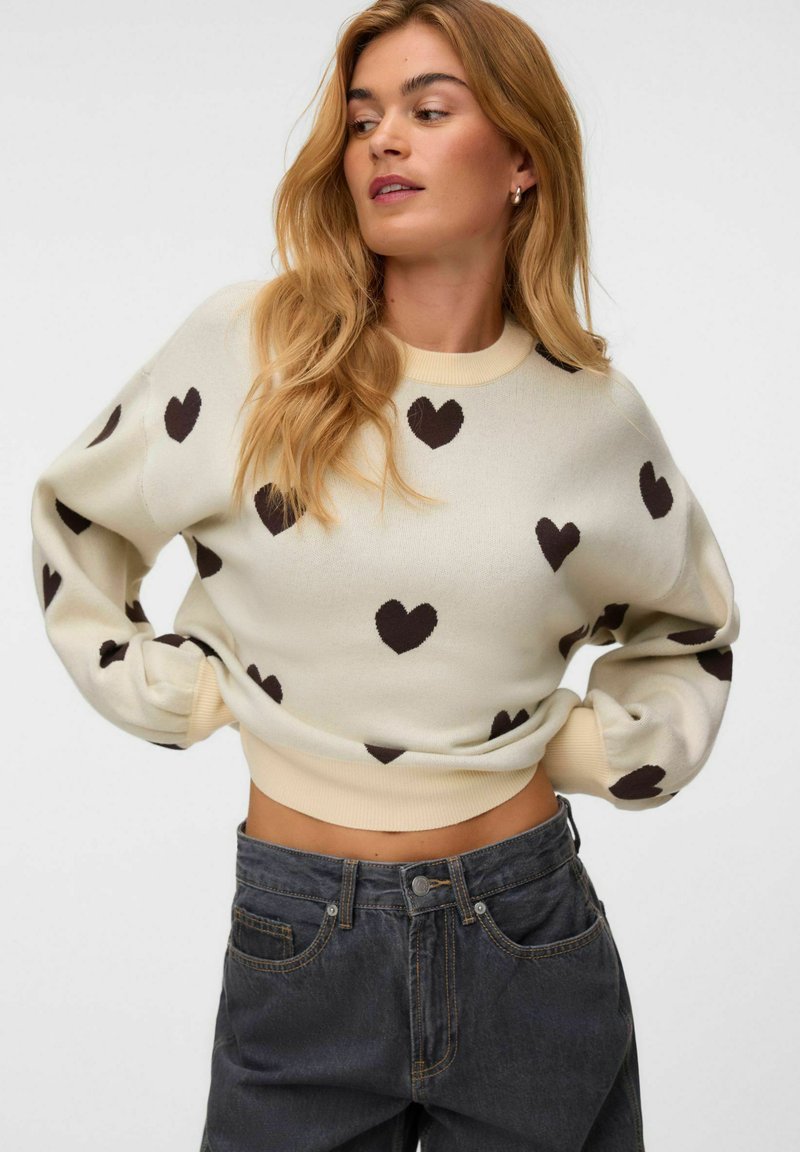 Femme aux longs cheveux blonds portant un pull crème avec des motifs de cœurs noirs et un jean taille haute gris foncé, posant les mains sur les hanches.