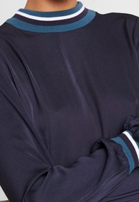 Maglietta a maniche lunghe blu navy con colletto a coste caratterizzato da righe blu e bianche. Tessuto morbido e leggero con polsini arricciati per un tocco di dettaglio.