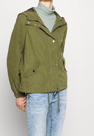 Persona con chaqueta con capucha de color verde oliva con botones automáticos sobre un jersey de cuello alto verde claro y vaqueros azul claro, manos relajadas a los lados.