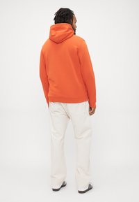 Uomo con dreadlocks che indossa un felpa arancione, pantaloni bianchi larghi e sneakers bianche con strisce nere, in piedi di fronte a un muro bianco.