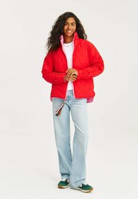 Červená prešívaná bunda s vysokým golierom, ktorá má ružovú podšívku a zips s multikolórovými strapcami. Nosená cez biele tričko a modré rifle.