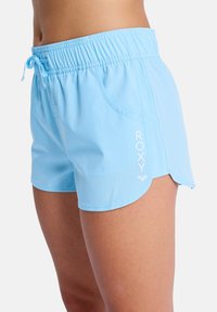 Woman wearing light blue elastic waist shorts with side pockets and "ROXY" logo printed on left leg.