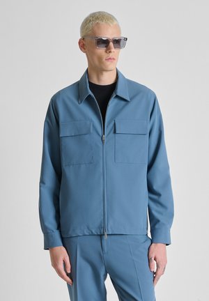 Jonge man met kort blond haar, die een rechthoekige zonnebril draagt en een blauw jack met rits en klepzakken, gecombineerd met bijpassende blauwe broek.