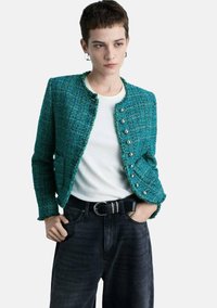 Giacca in tweed turchese con scollo rotondo, chiusura con bottoni frontali e maniche corte, abbinata a una maglietta bianca e jeans scuri.
