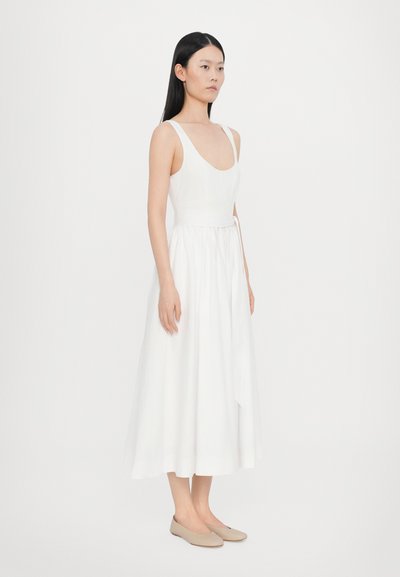 Weißes, ärmelloses Kleid mit weitem Rundhalsausschnitt und tailliertem Bund. Der fließende Stoff reicht bis zur Mitte der Wade und wird durch ein seitliches Bindeelement ergänzt.